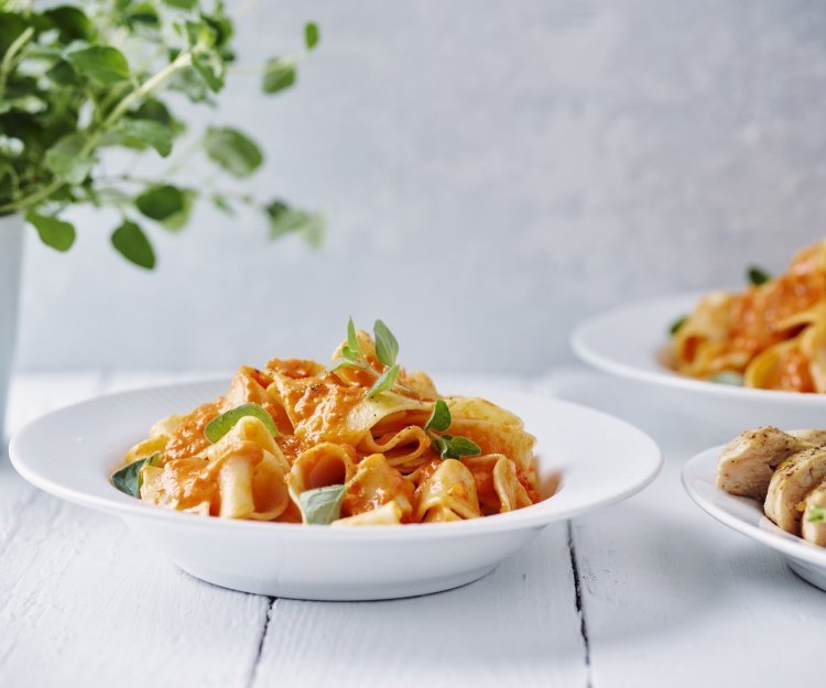 Pasta met geroosterde paprikasaus en kipfilet Een portie pasta met homemade paprikasaus staat klaar op een witte houten tafel. Een vers plantje oregano staat achteraan in beeld.