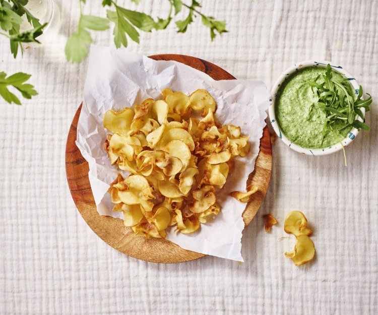 Op tafel ligt een kalkwit tafelkleed, met hierop een houten bordje met wit boterpapiertje en chips van pastinaak op. Er staat een dippotje langs met de dip van broccoli en roomkaas. 
