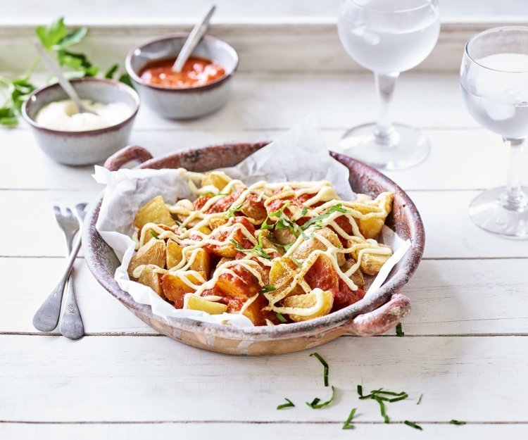 Heerlijke patatas bravas geserveerd in een groot diep bord, afgewerkt met een pittige tomatendip en aïoli. Twee gekoelde watertjes staan rechts van het bord. Klaar om de tapas tafel aan te vallen.