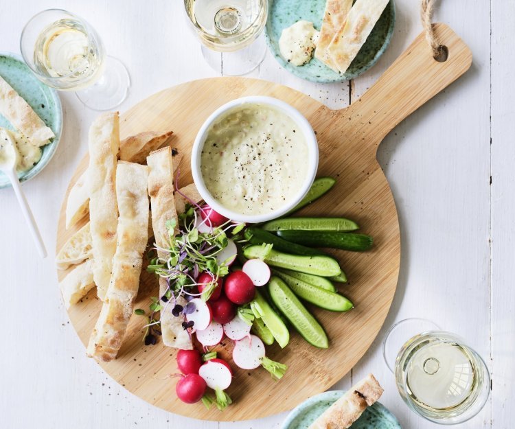 Pikante yoghurtdip met witte bonen en kruiden Op tafel staat een ronde houten snijplank met hierop een wit potje gevuld met onze pikante dip van yoghurt en witte bonen. Je kan de yoghurtdip gebruiken met stukjes snackkomkommer, brood en radijsjes in ons geval. Rond de plank staan enkele glazen witte wijn en helderblauwe broodbordjes.