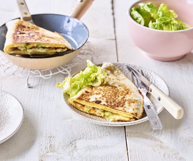 Een krokante tortilla ligt in een bordje klaar om aan te vallen. Hij is gevuld met een omelet, passendale kaas, gandaham en lente-ui. De tweede quesadilla ligt nog in de pan en staat ook op de tafel. 