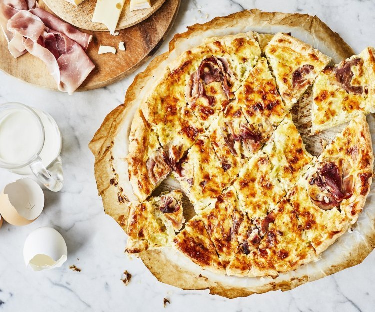 Quiche Lorraine Vers uit de oven staat deze quiche te schitteren op het marmere aanrecht. Hij is aangesneden in kleinere stukjes om aan te vallen tijdens het aperomoment. Er liggen enkele eitjes naast ter decoratie en verse boerenham.