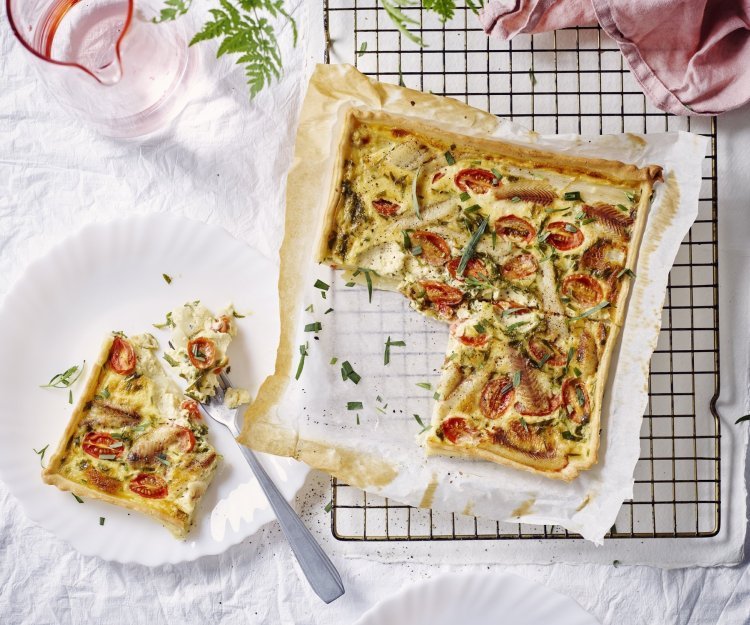 Vers gebakken uit de oven staat de quiche met asperges, tomaten en gerookte paling te blinken op tafel. Er werd al een stukje aangesneden en op een bordje gepresenteerd. Roze linnen servet en waterkaraf zorgen voor de sfeer in de foto. 