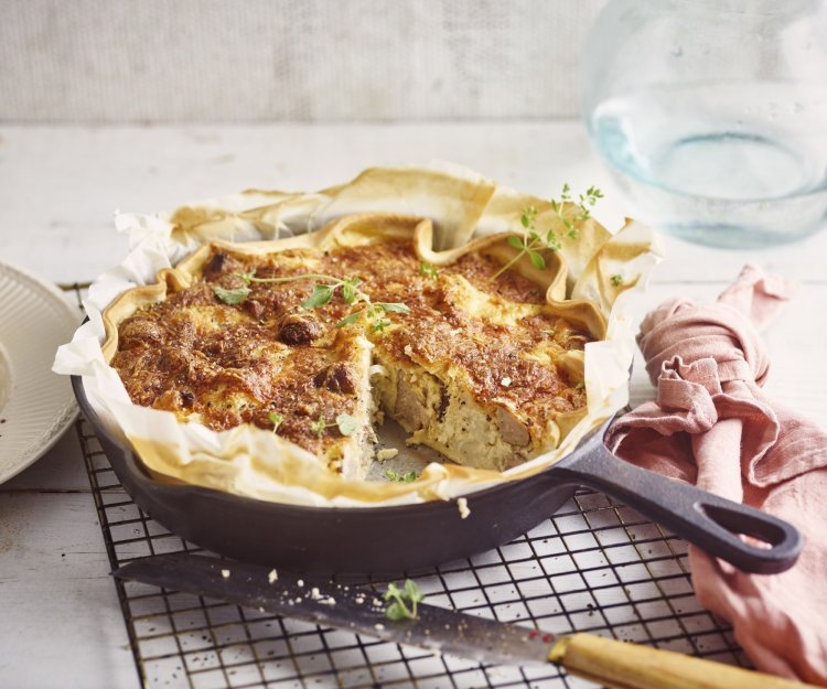 Quiche met bloemkool en worst Op het aanrecht ligt een zwarte rooster met hierop de quiche in een gietijzeren pan. Hierbij ligt een roze servet, mes en bordje waarop al een stukje quiche op ligt. De quiche is afgewerkt met enkele verse blaadjes oregano.