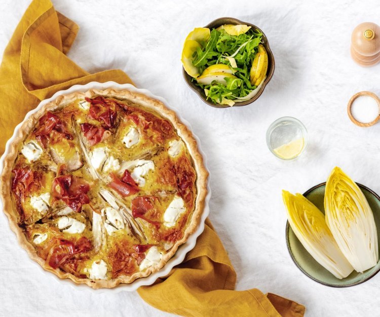 Quiche met witloof, peer en gedroogde ham Een ronde quiche met wiltoof staat op een okkerkleurige handdoek op het aanrecht.