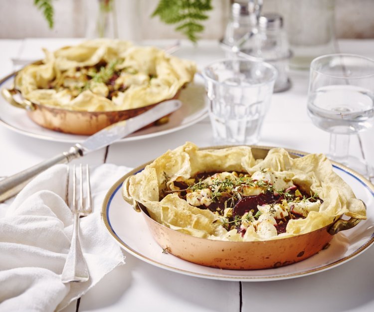 Aan tafel! Quiche van filodeeg met geitenkaas en rode biet staan klaar op tafel. De quiches zitten in een kopperen schaaltje en staan op een wit bord. Het bestek en de witte servette liggen langs het bord. Op tafel staan ook waterglazen en een glazen peper- & zoutvat.