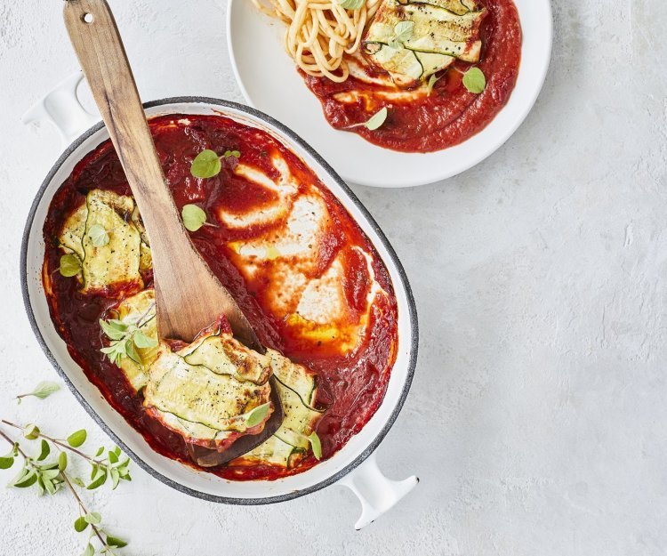 Pakjes van courgette liggen in witte ovenschaal met blauw randje. De tomatensaus ligt onder de ravioli van courgette, waardoor deze goed herkenbaar zijn. Er werd net een portie uitgeschept om meteen te eten. 