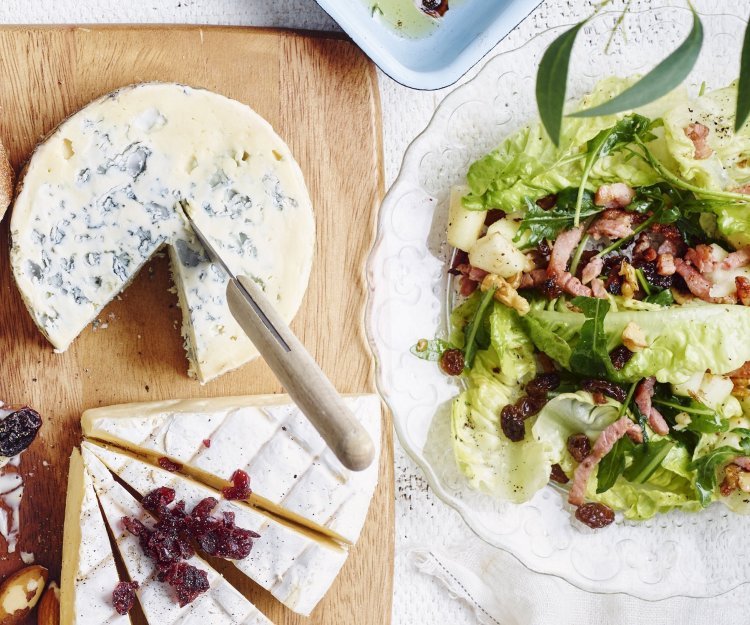 Een heerlijk salade met little gems, frisse appelstukjes en krokante spekblokjes staan op tafel in een glazen slakom. De slakom staat naast een kaasplank met een stuk blauwekaas en Belgische brie.