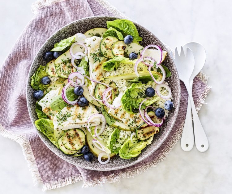 Een diep bord gevuld met sla, gegrilde zeetong, courgette, peer en blauwe bessen. Onder het bord ligt een paars servette en wit bestek om meteen dit gerechtje te verorberen. 