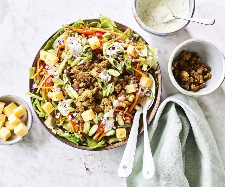 Salade met gehakt en ranchdressing Een grote salade kom staat op het aanrecht, hij is gevuld met gemengde sla, komkommer, tomaten, worteltjes, stukjes kaas en gebakken gehakt. We werkte de salade af met een ranchdressing op basis van zure room en verse kruiden.