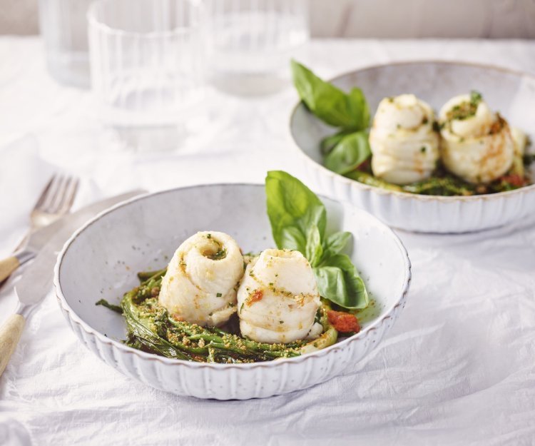 Schartongrolletjes met tomatenboter, zeekraal, lente-uitjes en crumble van ansjovis en peterselie Twee bordjes staan op tafel, het zijn diepe borden in een lichtblauwe/grijze kleur. Ze zijn gevuld met de lente-uitjes en zeekraal op de bodem in een nestjesvorm. Hier bovenop liggen de schartongrolletjes. Het gerechtje is afgewerkt met de tomatenboter en crumble van ansjovis en peterselie.