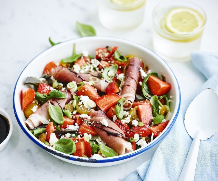 Rijkgevulde salade met aardbeien, geitenkaas en gandaham. Afgewerkt met balsamico. Er zijn twee frisse waters boven de kom met elk een citroentschijfje in. 