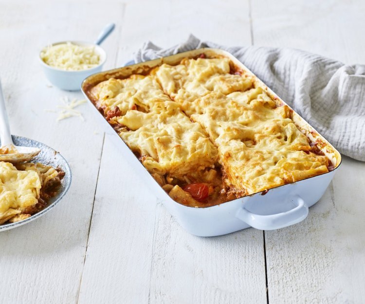 Slowcooked mac & cheese lasagne Een lichtblauwe ovenschotel staat op een houten witte tafel. De ovenschotel is gevuld met een mac & cheese én een tomatensaus, waardoor hij eerder weg heeft van een lasagne. Er is al 1 portie uitgeschept en wil al beginnen aan de volgende. Laat het niet koud worden.