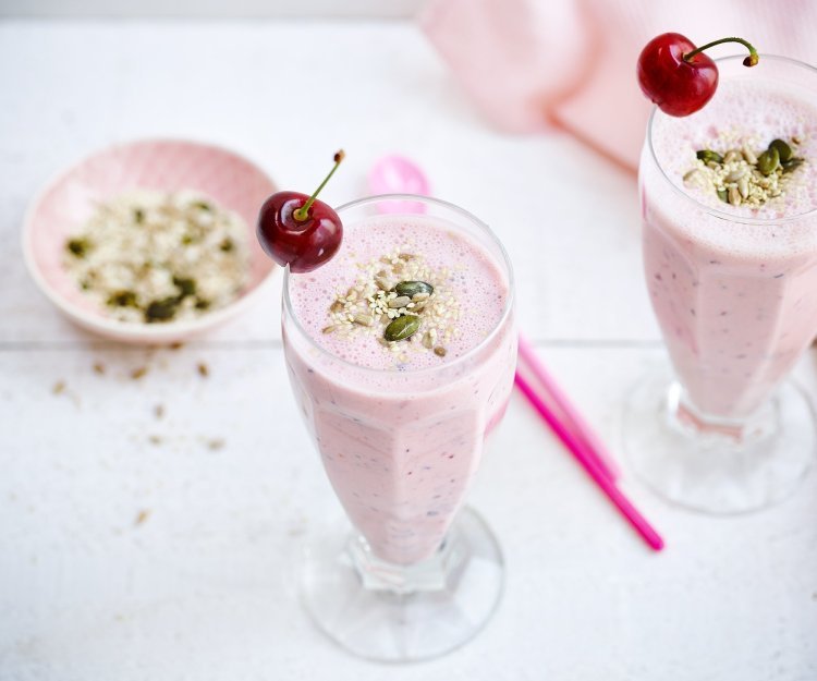 Smoothie van kersen en blauwe bessen Twee rooskleurige smoothies op een witte houten tafel. De smoothie is afgewerkt met een kers en enkele pitjes en zaadjes. Een snel en makkelijk drankje met feestallures!