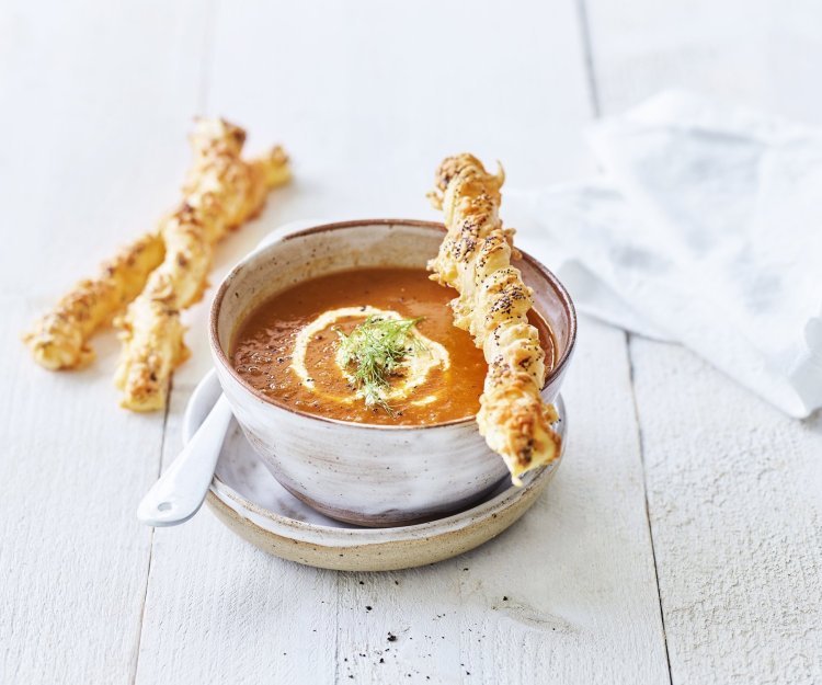 Een heerlijke kop soep staat te pronken op de eettafel. Het is een rood-oranje kleurige soep waarbij enkele bladerdeegstukjes liggen gevuld met kaas. De soep is afgewerkt met een lepeltje room en een peterselieblaadje. 