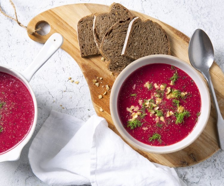 Soep van rode biet met aardappel, zure room en komijn Soep van rode biet kleurt natuurlijk rood en smaakt naar nog! Ze is geserveerd in twee witte kommetjes en staat op een houten snijplank met enkele bruine sneden brood bij.