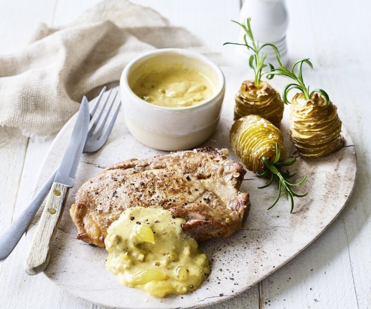 Een bordje om meteen aan te vallen! Een sappige varkenskotelet met picklessaus en aardappeltorentjes met verse rozemarijn. Het bestek ligt langs het bordje, bovenaan een witte pepermolen en servette.