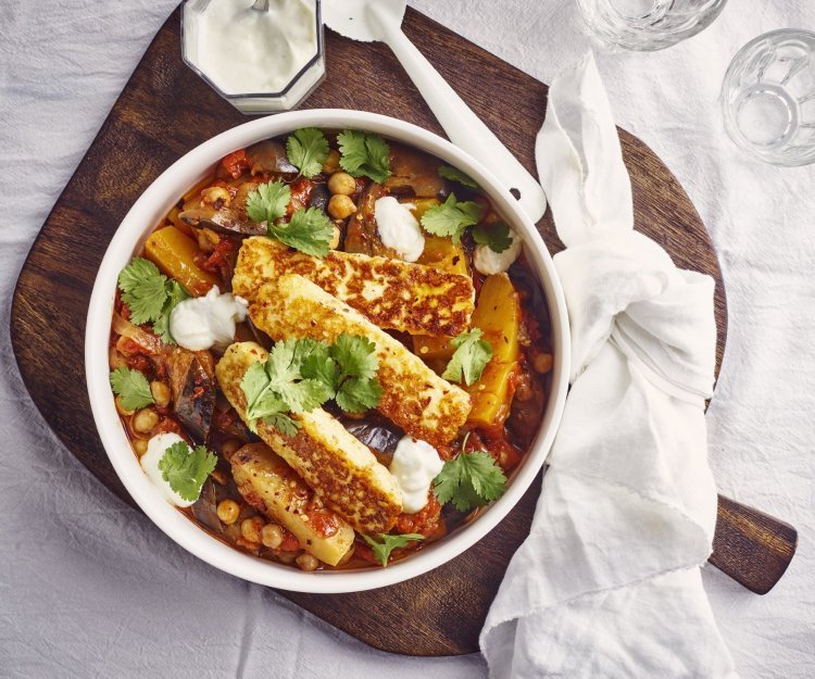 Stoofpot van aubergine en pompoen met Berloumi Op tafel ligt een wit tafelkleed met hierop een donker houte plank. Hierop staat het stoofpotje met een witte geknooptje handdoek langs. Er staat een glaasje langs met knoflookyoghurt en twee waterglazen. Het groentestoofpotje heeft een mooie warme kleur door de oranje pompoen, groene korianderblaadjes en de gegrilde kaas.