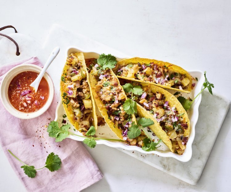 Taco’s met aardappelen en gehakt Vier tacos liggen klaar in een ovenschaal om aan te vallen. De pittige tomatendip staat links van de schotel op een roze servette. Het gerechtje is afgewerkt met verse korianderblaadjes.