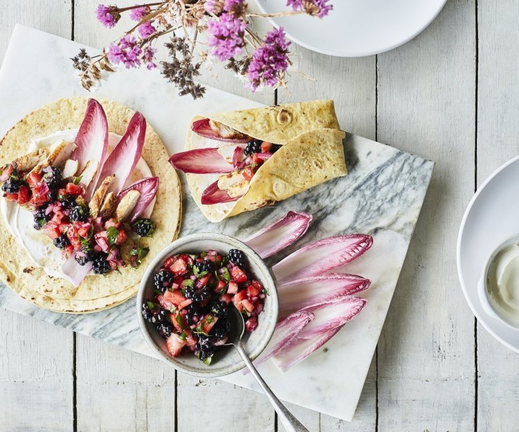 Taco’s met kip, roodlof en een salsa van braambessen en aardbei ligt klaar om op te eten. Er ligt al een tweede taco in de maak, op een marmere snijplank. De kleuren van roos tot donker rood van het fruit en loof geven een mooi contrast in de foto.