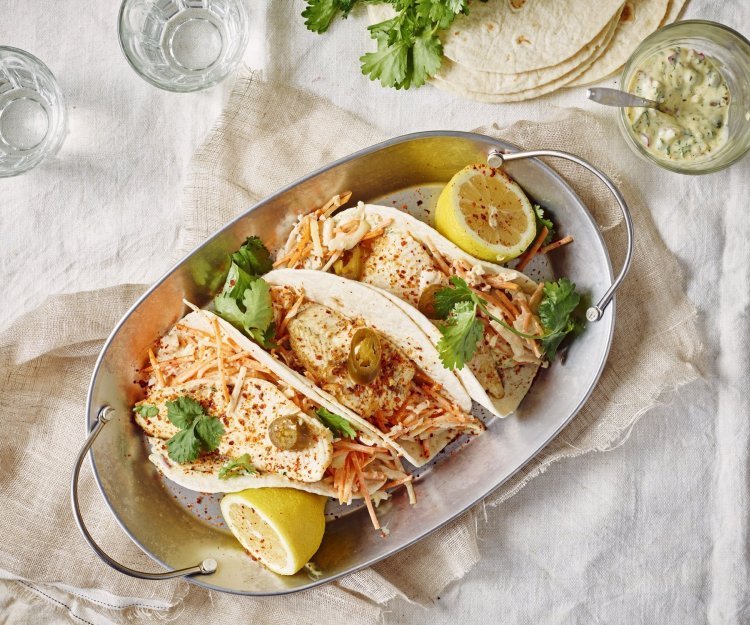 Taco’s met pladijs, koolsla en tartaarsaus Drie wraps liggen gepresenteerd in een ovenschotel van inox. Deze staat op een gedekte tafel met wit linnen kleed. Er liggen twee citroenhelften bij de tortilla's en verse koriander.