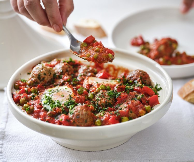 Tajine met gehaktballetjes, tomatensaus en ei Op tafel staat een witte tajine heerlijk gevuld met gehaktballen in een rijke tomatensaus met eitjes. Er wordt net een portie uitgeschept.