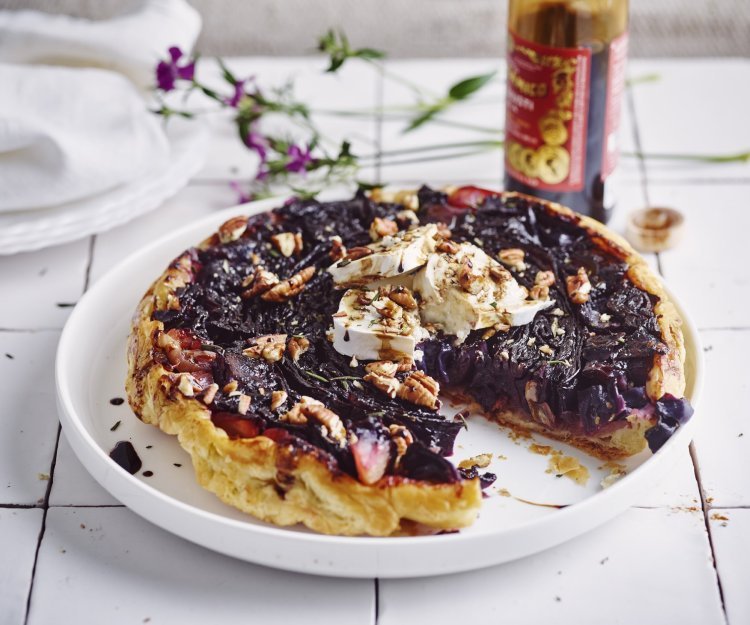 Een tarte tatin van rode kool met geitenkaas ligt op een groot wit bord. Er is al een stukje aangesneden en ze werd afgewerkt met enkele druppels balsamico en pecannoten. Achteraan zie je de fles balsamico nog op het aanrecht staan alsook een paars bloemetje. 