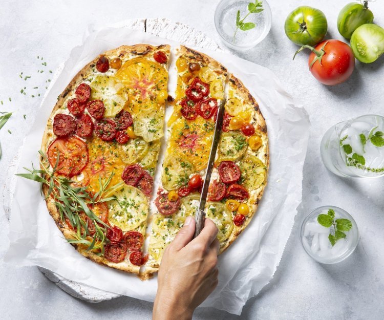 Tomatentaart met roomkaas en verse kruiden Een kleurrijke tomatentaart staat op tafel op een wit boterpapiertje. Ze wordt aangesneden door een onbekende vrouw met een koksmes. Rechtsboven liggen nog enkele tomaten om ze extra in de picture te zetten.