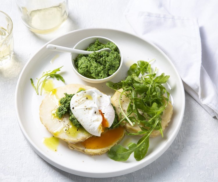 Traaggegaarde knolselderschijven liggen op een groot wit bord vergezeld met een gepocheerd ei en pesto van boerenkool in een wit potje. Er is ook nog wat rucola toegevoegd voor de extra groene toets.   