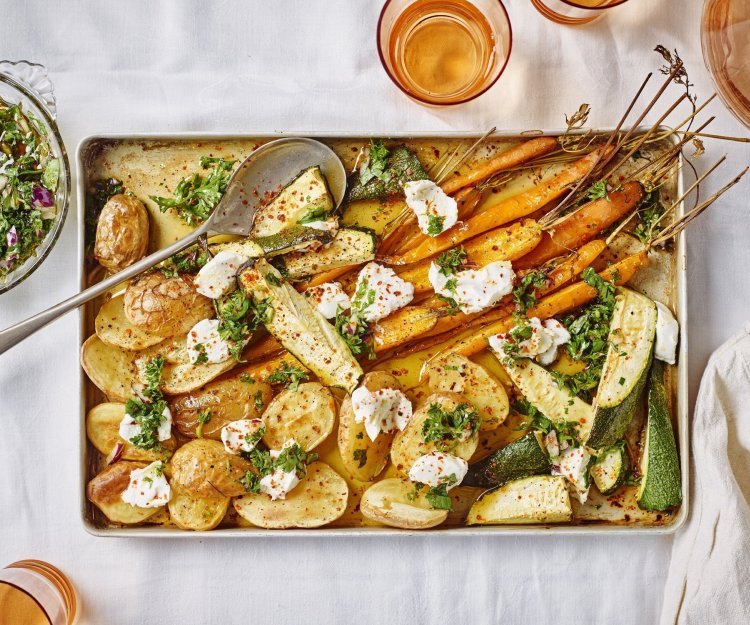 Ovenplaat met gegrilde groentjes komt net uit de oven en staat al op tafel. Hij heeft warme kleuren van de gouden aardappelen naar de oranje wortelen. Hij is afgewerkt met toefjes geitenkaas en verse chimichurri.