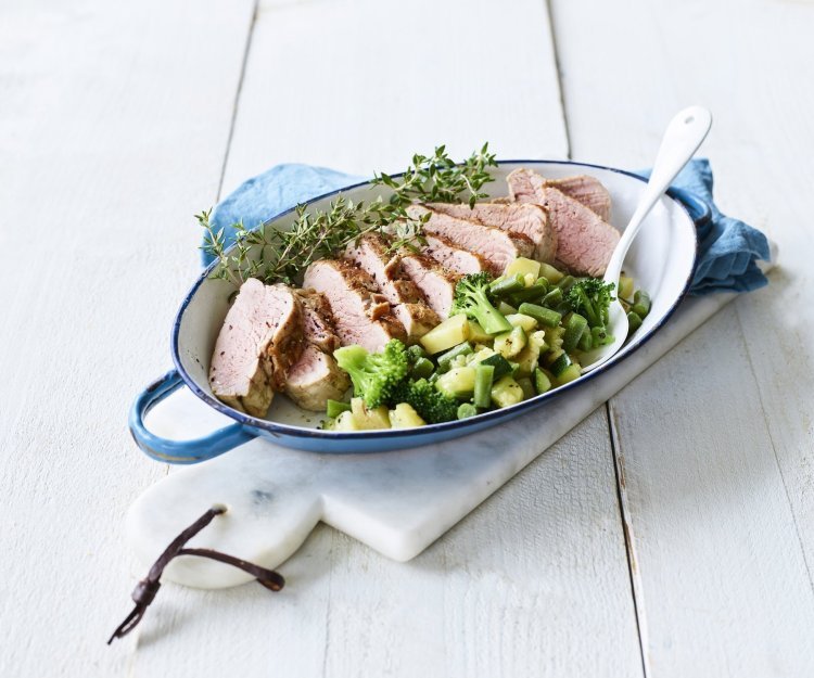 Een heerlijk varkenshaasje uit de oven ligt in een blauwe ovenschaal, vergezeld met groene groenten en verse tijm. 