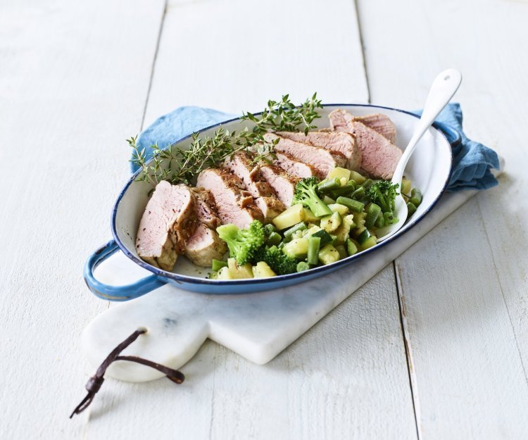 Varkenshaasje in de oven met macedoine van groene groenten Een heerlijk varkenshaasje uit de oven ligt in een blauwe ovenschaal, vergezeld met groene groenten en verse tijm.