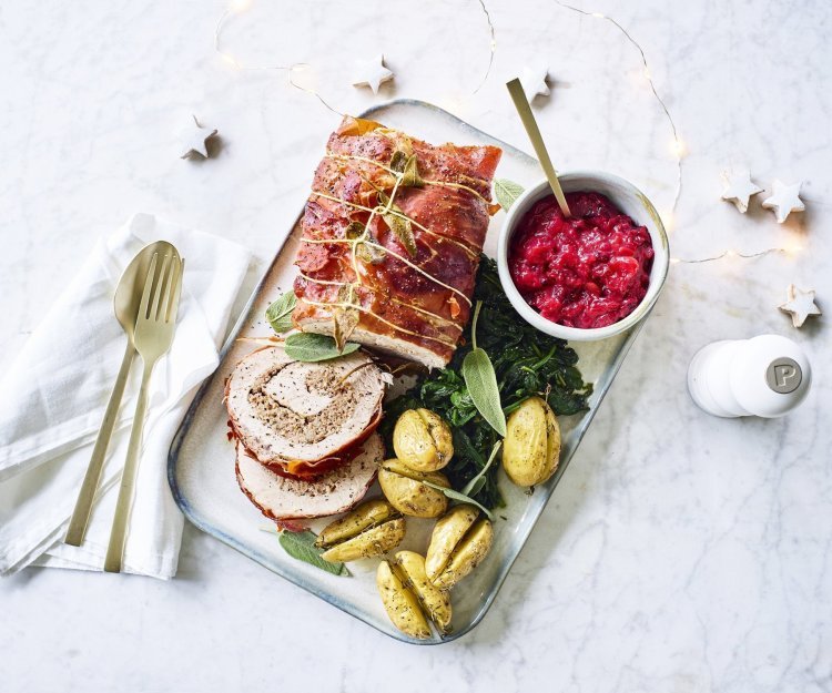 Varkensrollade met gehakt, veenbessencompote en krieltjes uit de oven Een mooi aangesneden varkensrollade ligt op een rechthoekig dienblad. De aardappeltjes werden net niet doormidden gesneden alvorens ze de oven in gingen met een kruidentakje. De veenbessencompote staat langs de rollade die zorgt voor een intense kleur in deze feestmaaltijd. Goud bestek ligt langs het gerecht.