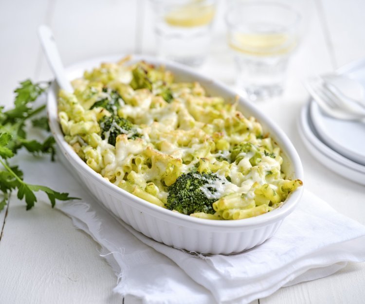 Vegetarische Mac'n cheese met witte bloemkoolsaus en broccoli Een grote ovenschaal staat op een houte witte tafel. Hij staat op een wit lapje stof. De ovenschotel is gevuld met macaroni, broccolie en witte bloemkoolsaus. Er staan enkele bordjes langs met wit bestek om direct aan te vallen.