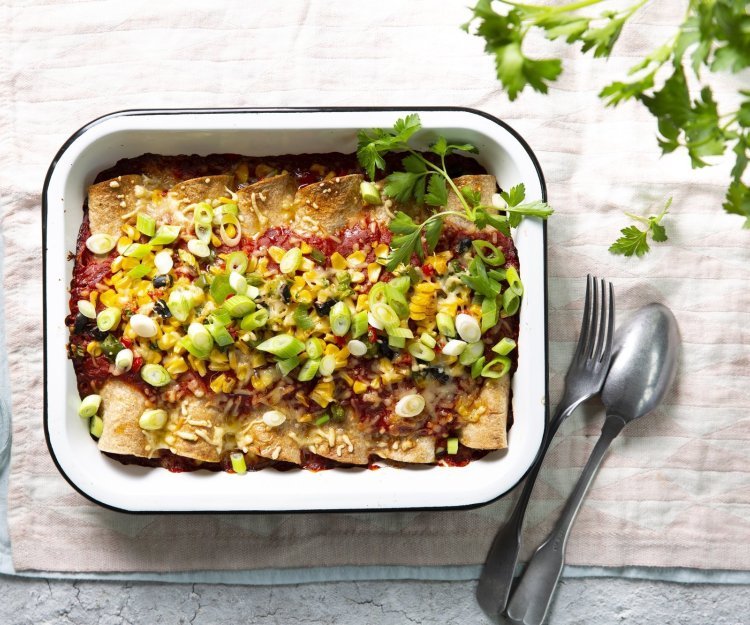 Enchiladas liggen mooi op een rijtje in een witte ovenschotel. Hierop werd tomatensaus geserveerd en gemalen kaas, in de oven. Versgesneden lente-uitjes werden als afwerking toegevoegd aan het gerecht. 