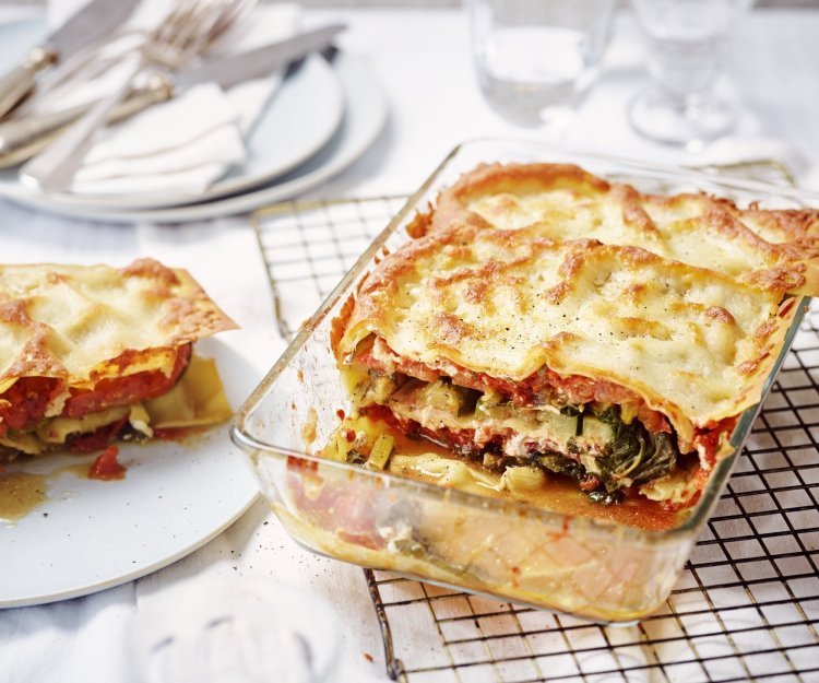 Vegetarische lasagne met snijbiet, courgette en ricotta De lasagne staat op tafel in een glazen doorzichtige ovenschotel. Hij is aangesneden zodat je mooi de laagjes ziet van pasta, groenten en saus. De portie die is uitgeschept ligt op een bordje er naast.