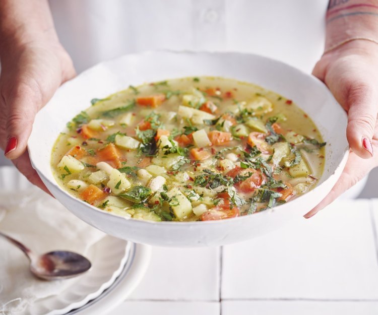 Een vegetarische minestrone met venkel, witte bonen, aardappel en verse kruiden geserveerd in een diep wit soepbord. Een vrouw met mooi rood gelakte nagels draagt het bord. 