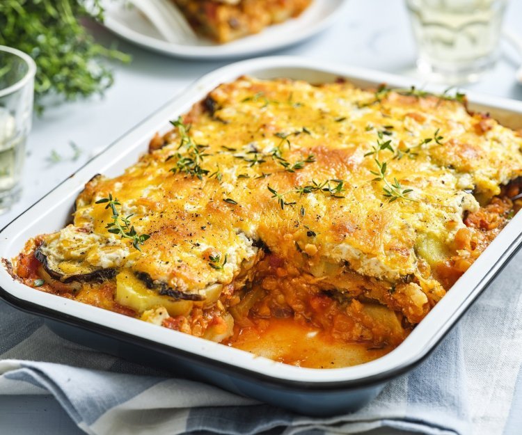 Er staat een grote schotel met vegetarische moussaka op tafel. Er is al een portie uitgeschept en op een bordje geserveerd. De moussaka is afgewerkt met verse tijmblaadjes. Op tafel staan ook nog 2 glazen en het plantje verse tijm.