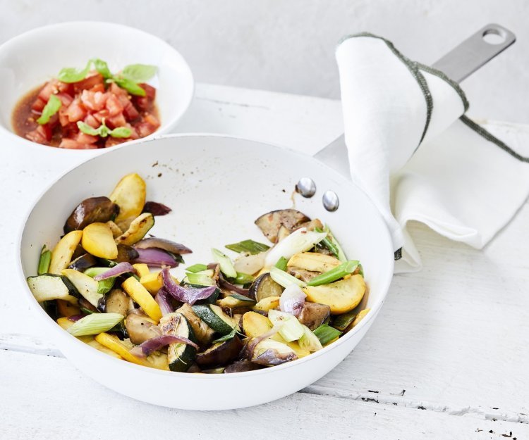 Vegetarische wok van aubergine en courgette staat klaar om aan te vallen in een witte wokpan. Er is een potje gevuld met tomatenblokjes, basilicum, azijn en olijfolie waarmee je je gerecht een extra frisse toets geeft.