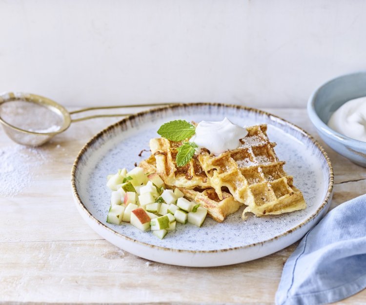 Verloren brood-wafeltjes met appel & peer Wafeltjes van verloren brood liggen op een licht grijs bordje. De stukjes fruit van appel en peer liggen onder de wafeltjes. We gaven ook een toefje slagroom op de wafels voor extra smaak. Er ligt een blauwe servette en klein zeefje met bloemsuiker langs het bord.