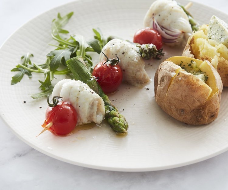 Een spies van visrolletjes, groene asperges en kerstomaten ligt op een wit bord. Ze worden geserveerd met een goudgele gepofte aardappel en natuurlijk een klompje boter erin. 