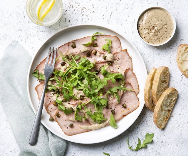 Deze vitello sgombro ligt op een wit bord, heerlijk afgewerkt met de koude saus, kappertjes en rucola. Er liggen ook enkele stukjes brood langs het bord.