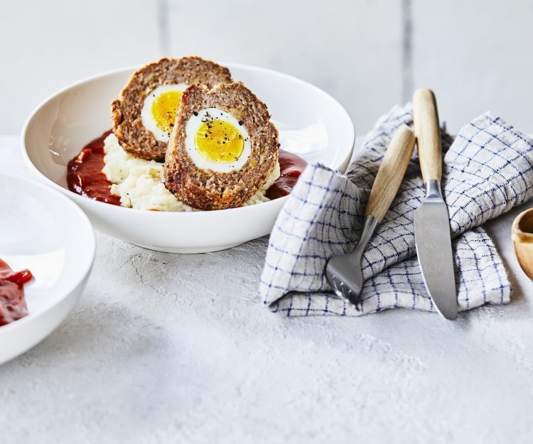 Vogelnestjes in tomatensaus met aardappelpuree In een diep wit bord ligt de aardappelpuree onderaan met verse rode tomatensaus en daar bovenop de doormidden gesneden vogelnestjes. Er ligt een handdoek langs met blauwe ruitjes, hierop het bestek met houten handvat.