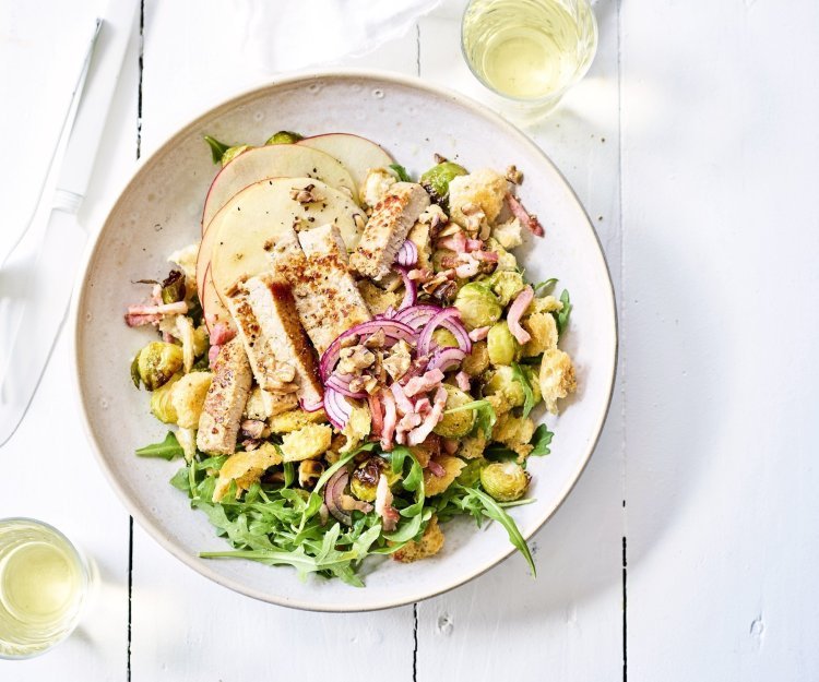 Winterse panzanella met ovengeroosterde spruitjes, versgesneden appel en gegrilde kalfsoester in een diep wit bord. Wit bestek en wijn staat langs het bord, klaar om aan te vallen.