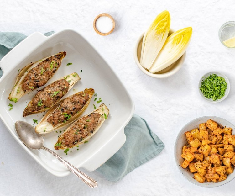 Witloof gevuld met gehakt en champignons met kruidige aardappelen Gevulde kroppen witloof met kruidig gehakt liggen in een ovenschotel. De schotel staat op een lichtblauwe doek met kruidige aardappelen in kommetje langs.