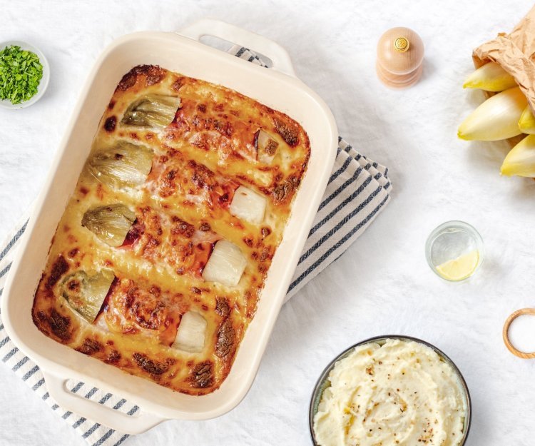 Gegratineerde witloofrolletjes opgerold in gekookte ham liggen mooi naast elkaar in een ovenschotel. De kaassaus is laat de rolletjes net bovenkomen. Er staat een pot heerlijke puree klaar om aan te vallen. 