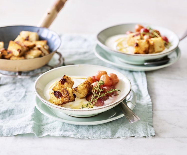 Een heerlijk én makkelijk ontbijtje staat klaar op tafel. De yoghurt is geserveerd in ontbijtkommetjes en afgewerkt met gegrilde druiven en krokante croutons van rozijnenbrood.