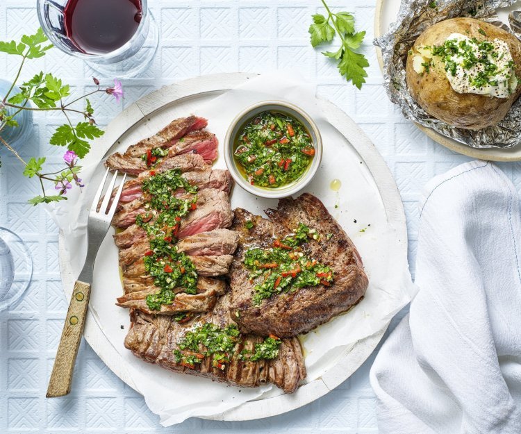 Het heerlijk stukje vlees ligt op een groot wit rond bord. Het al voor de helf in reepjes gesneden en bedruppeld met de Aziatische chimichurri. Er ligt een gepofte aardappel langs en twee glaasjes rode wijn. 