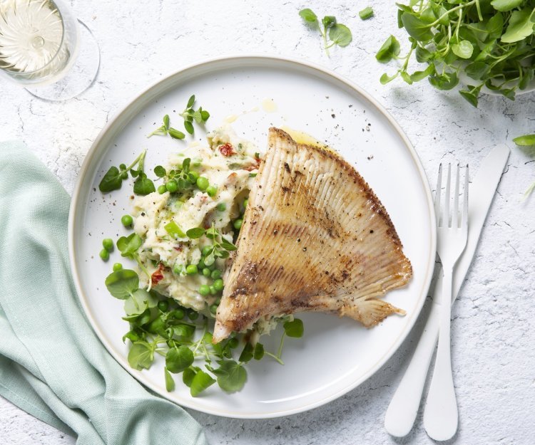 Zuiderse puree met rog Een wit bord gevuld met een frisse schep van de puree boordevol extra groentjes, denk maar aan verse lente-uitjes, zongedroogde tomaatjes, waterkers, kruiden enz. Hierop werd de gebakken rogvleugel gelegd die mooi krokant is gebakken. Naast het bord ligt ook nog een lichtblauwe servette, kommetje met extra waterkers en een glaasje wijn. Smakelijk!