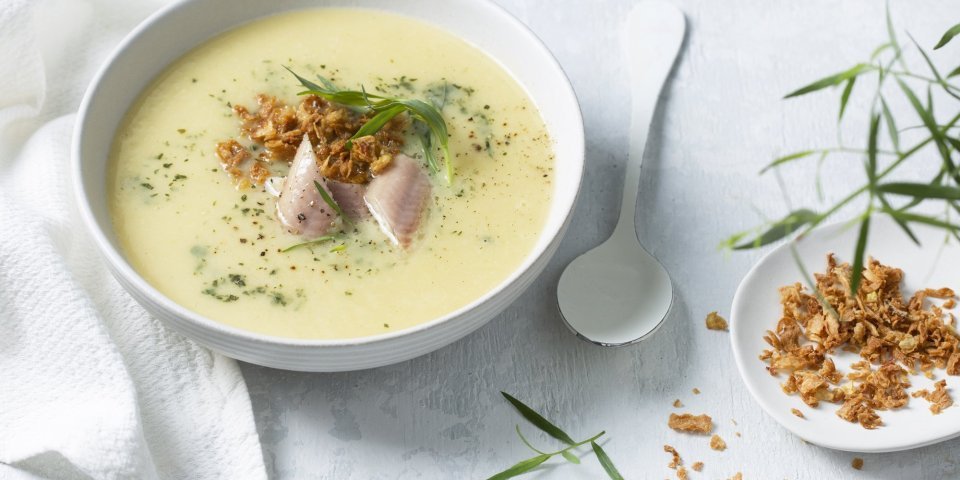 Aardappelsoep met gerookte paling en krokante uitjes Dampende aardappelsoep met stukjes paling in het midden. We geven ze extra kleur en smaak door dragon toe te voegen en krokante uitjes. Langs het bord ligt een witte eetlepel en extra uitjes en dragon.